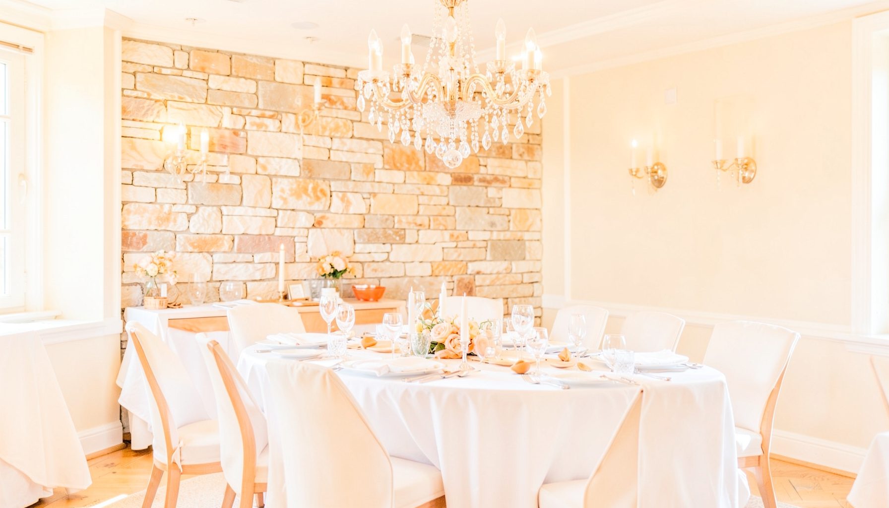 Bright, airy photograph of an elegant dining room with crystal chandelier, stone accent wall, wall sconces, and white tablecloths