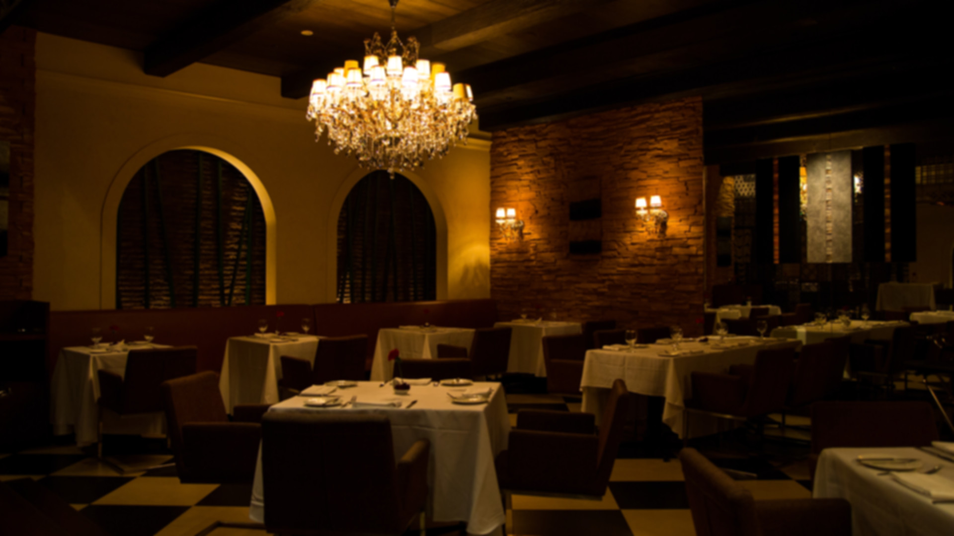 Elegant restaurant dining room with crystal chandelier, stone accent wall, wall sconces, white tablecloths, and checkered flooring